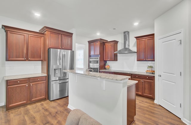 kitchen featuring a center island with sink, wall chimney range hood, appliances with stainless steel finishes, light stone counters, and wood-type flooring