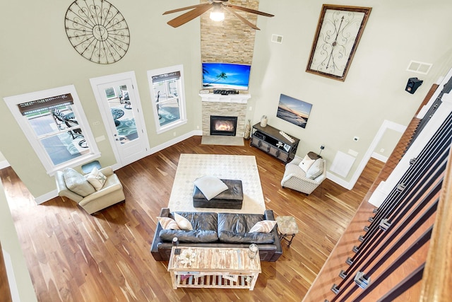 living room featuring ceiling fan, a towering ceiling, wood-type flooring, and a stone fireplace