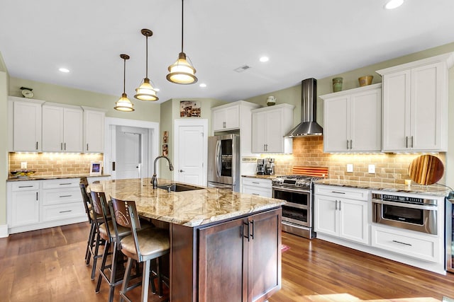 kitchen featuring stainless steel appliances, white cabinetry, decorative light fixtures, an island with sink, and wall chimney exhaust hood