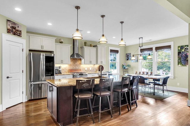 kitchen featuring stainless steel appliances, a center island with sink, white cabinetry, light stone counters, and wall chimney range hood