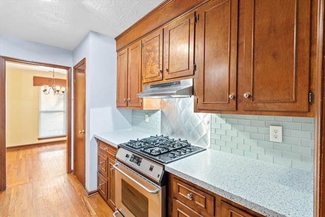 kitchen with brown cabinets, a notable chandelier, light wood-style flooring, under cabinet range hood, and stainless steel range with gas stovetop