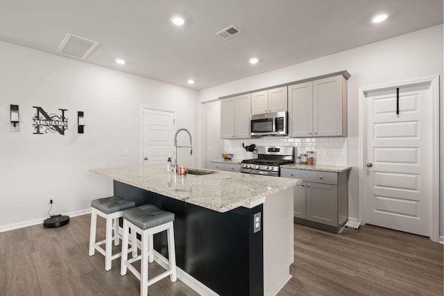 kitchen featuring appliances with stainless steel finishes, a sink, gray cabinetry, dark wood-style flooring, and a center island with sink