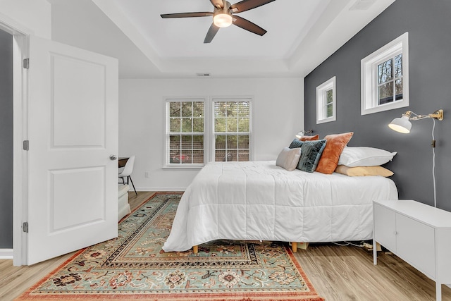 bedroom with ceiling fan, visible vents, baseboards, light wood-style floors, and a tray ceiling