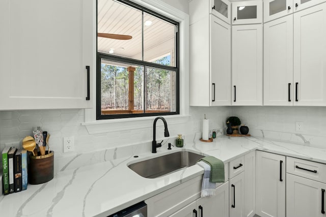 kitchen featuring a sink, white cabinets, backsplash, light stone countertops, and glass insert cabinets