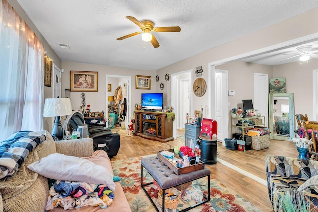 living room with ceiling fan, a textured ceiling, wood finished floors, visible vents, and a glass covered fireplace