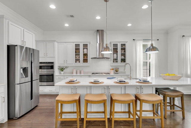 kitchen with appliances with stainless steel finishes, wall chimney range hood, a center island with sink, decorative light fixtures, and white cabinets