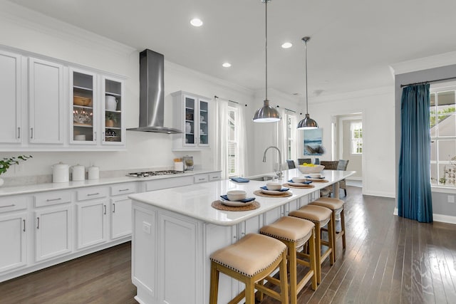 kitchen with a kitchen island with sink, white cabinets, decorative light fixtures, and wall chimney range hood