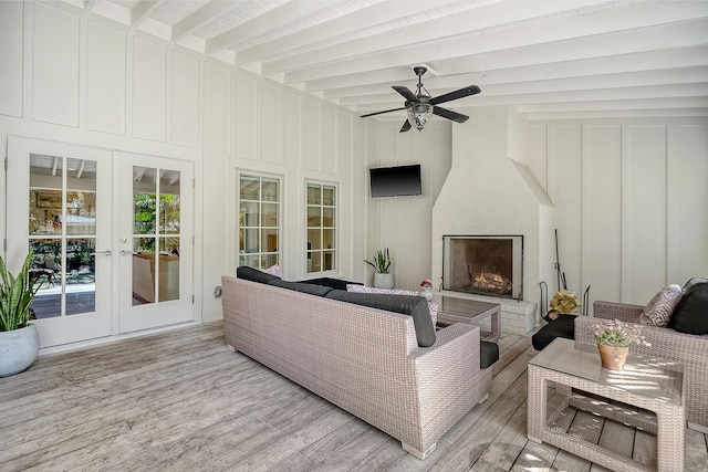 living area with a ceiling fan, light wood finished floors, lofted ceiling with beams, a warm lit fireplace, and french doors