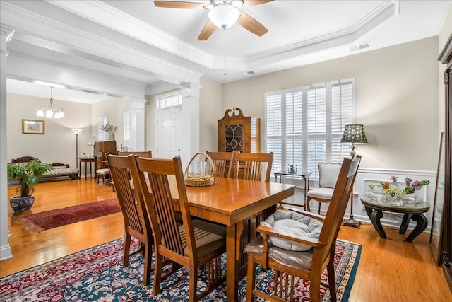 dining space with ceiling fan with notable chandelier, a raised ceiling, crown molding, light hardwood / wood-style flooring, and ornate columns