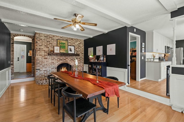 dining area featuring beamed ceiling, a wainscoted wall, light wood-style flooring, a fireplace, and a textured ceiling