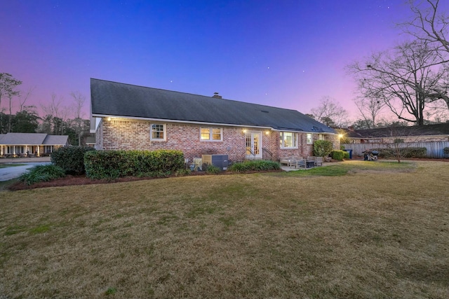 back of property at dusk with fence, a yard, brick siding, central AC unit, and a chimney