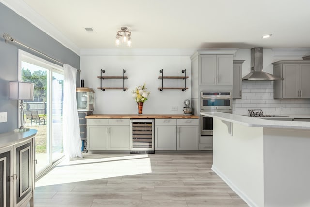 kitchen with wooden counters, wall chimney range hood, beverage cooler, gray cabinets, and stainless steel double oven