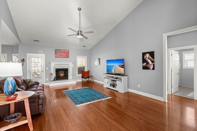 living room featuring wood finished floors, baseboards, high vaulted ceiling, ceiling fan, and a glass covered fireplace