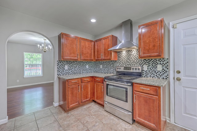 kitchen featuring wall chimney exhaust hood, stainless steel electric stove, backsplash, arched walkways, and a chandelier