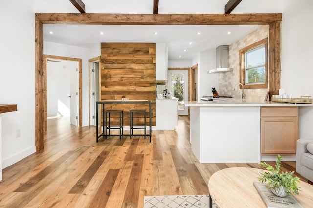 kitchen with wall chimney exhaust hood, light brown cabinets, beamed ceiling, light hardwood / wood-style floors, and decorative backsplash