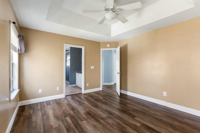 unfurnished bedroom with a tray ceiling, dark wood-style flooring, ensuite bath, and baseboards