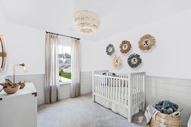 carpeted bedroom featuring a crib, wooden walls, a notable chandelier, and a wainscoted wall