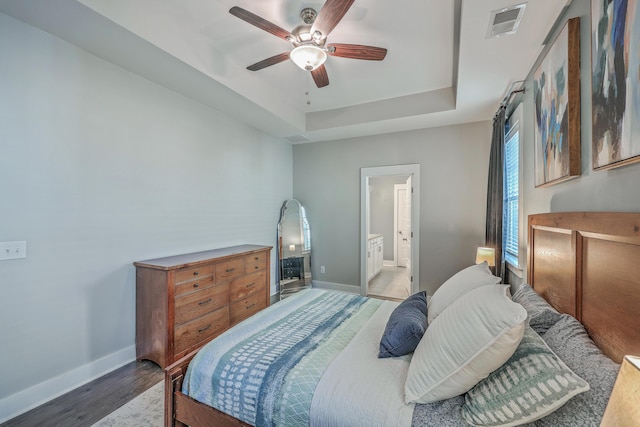 bedroom featuring visible vents, a raised ceiling, ensuite bathroom, wood finished floors, and baseboards