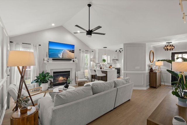 living room with a wainscoted wall, lofted ceiling, ceiling fan, a glass covered fireplace, and light wood-type flooring