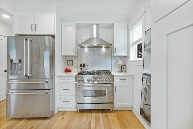 kitchen with light wood-type flooring, tasteful backsplash, stainless steel appliances, wall chimney range hood, and white cabinets