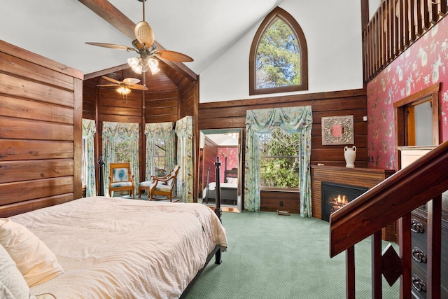 carpeted bedroom featuring wood walls, high vaulted ceiling, a lit fireplace, and a ceiling fan