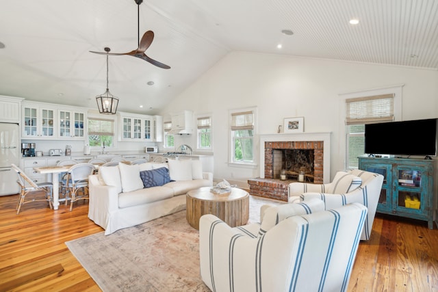 living room featuring light hardwood / wood-style floors, sink, ceiling fan with notable chandelier, high vaulted ceiling, and a brick fireplace