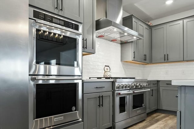 kitchen with light hardwood / wood-style flooring, wall chimney exhaust hood, gray cabinets, decorative backsplash, and stainless steel appliances