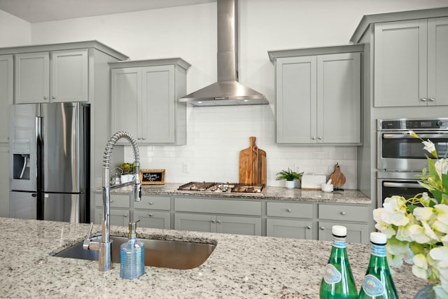 kitchen featuring tasteful backsplash, stainless steel appliances, gray cabinetry, wall chimney range hood, and a sink