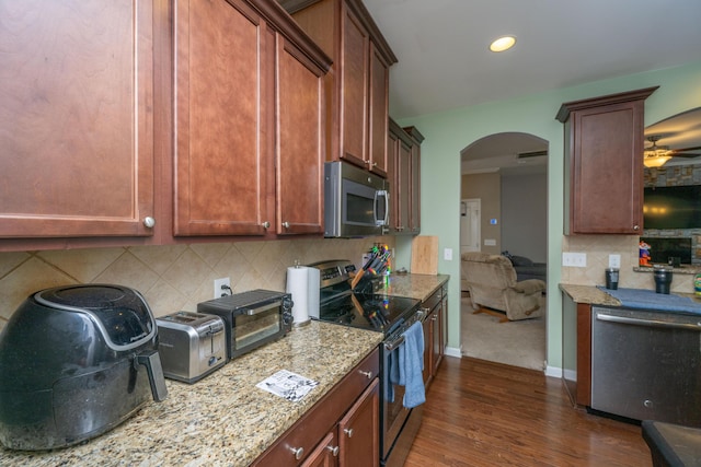 kitchen with dark wood-type flooring, backsplash, arched walkways, appliances with stainless steel finishes, and light stone countertops