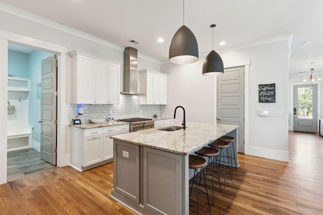 kitchen with wall chimney exhaust hood, sink, decorative light fixtures, white cabinetry, and a kitchen island with sink