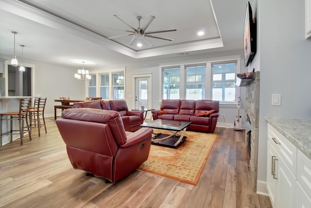 living room featuring baseboards, a tray ceiling, light wood-style flooring, ornamental molding, and ceiling fan with notable chandelier