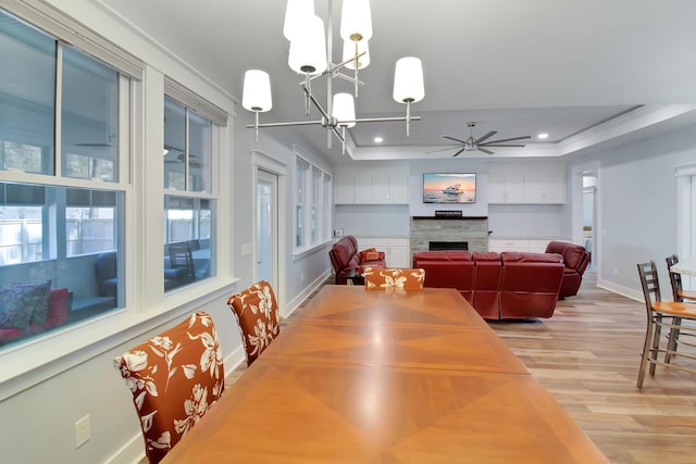 dining room featuring baseboards, light wood finished floors, a tray ceiling, a fireplace, and ceiling fan with notable chandelier