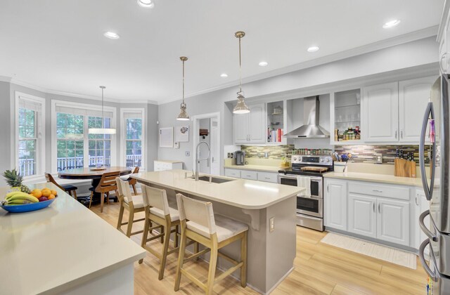 kitchen featuring appliances with stainless steel finishes, wall chimney exhaust hood, a sink, ornamental molding, and an island with sink