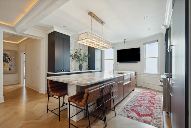 kitchen with a kitchen island with sink, a decorative wall, a sink, hanging light fixtures, and crown molding