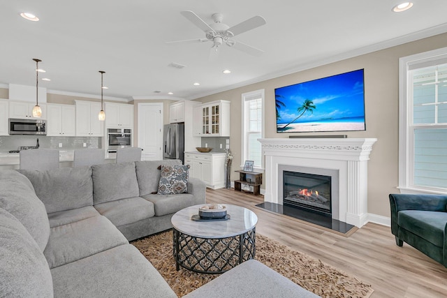 living room with ornamental molding, a glass covered fireplace, ceiling fan, and light wood finished floors