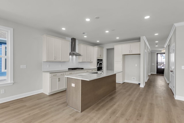 kitchen with wall chimney range hood, light wood-style flooring, an island with sink, and stainless steel gas stovetop