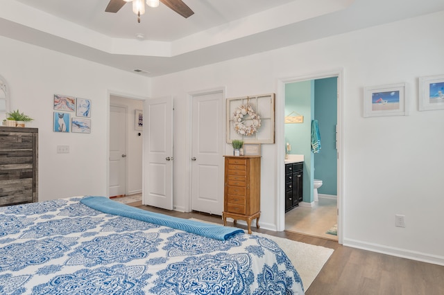 bedroom with ensuite bath, wood-type flooring, a raised ceiling, and ceiling fan