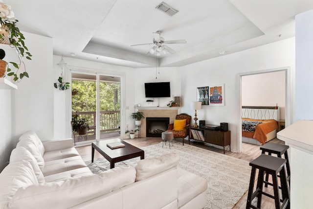 living room with a raised ceiling, a fireplace, a ceiling fan, light wood-type flooring, and visible vents