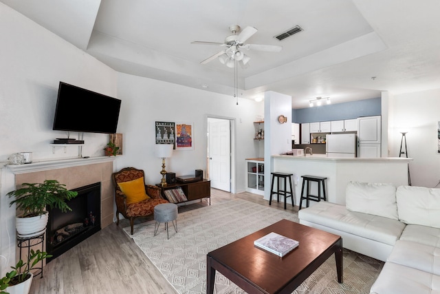 living area with light wood-style flooring, a ceiling fan, a tile fireplace, a tray ceiling, and visible vents