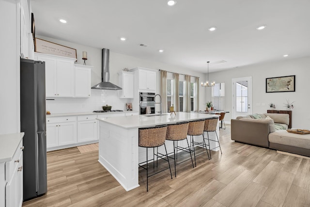 kitchen featuring wall chimney range hood, appliances with stainless steel finishes, an inviting chandelier, a kitchen breakfast bar, and open floor plan