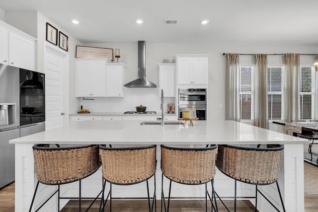 kitchen featuring visible vents, an island with sink, wall chimney range hood, light countertops, and stainless steel appliances