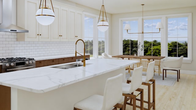 kitchen with light stone countertops, light hardwood / wood-style floors, wall chimney exhaust hood, stainless steel range, and a kitchen island with sink