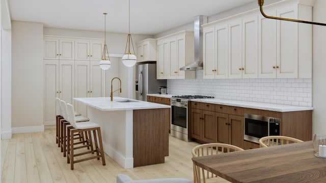 kitchen with wall chimney range hood, an island with sink, sink, appliances with stainless steel finishes, and light hardwood / wood-style floors