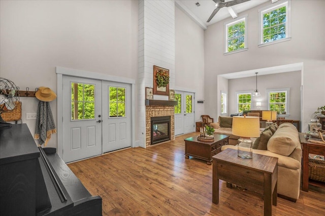 living area featuring a towering ceiling, a fireplace, wood finished floors, ceiling fan, and french doors