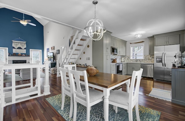 dining room featuring ceiling fan with notable chandelier, dark wood-style floors, a fireplace, stairs, and vaulted ceiling