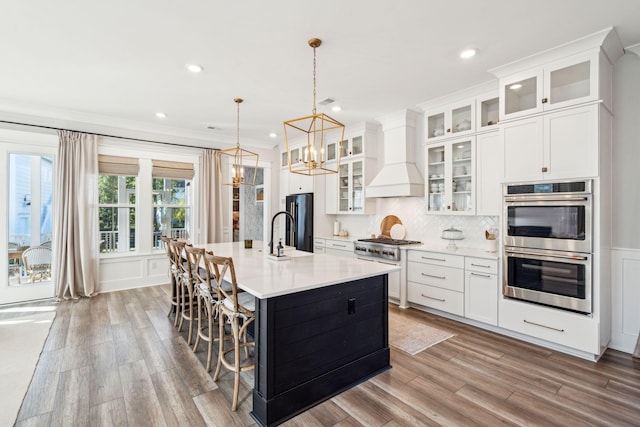 kitchen with appliances with stainless steel finishes, hanging light fixtures, light hardwood / wood-style flooring, premium range hood, and a center island with sink