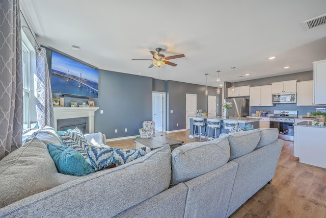 living area with light wood finished floors, visible vents, a glass covered fireplace, ceiling fan, and baseboards