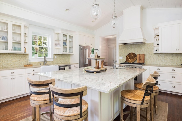 kitchen featuring white cabinetry, decorative light fixtures, custom range hood, stainless steel appliances, and a kitchen island with sink