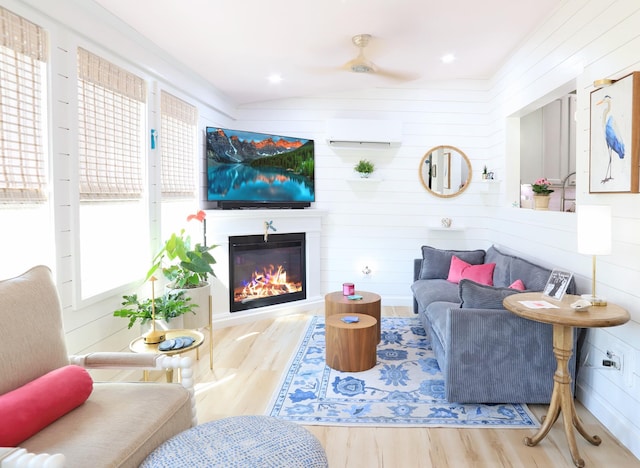 living room featuring wood finished floors, a ceiling fan, vaulted ceiling, a wall mounted AC, and a glass covered fireplace