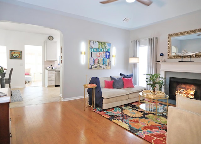 living area with arched walkways, ceiling fan, visible vents, light wood-style floors, and a lit fireplace